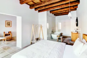 Moderní ložnice spojená s koupelnou, pokoj má trámový strop a kamennou podlahu.